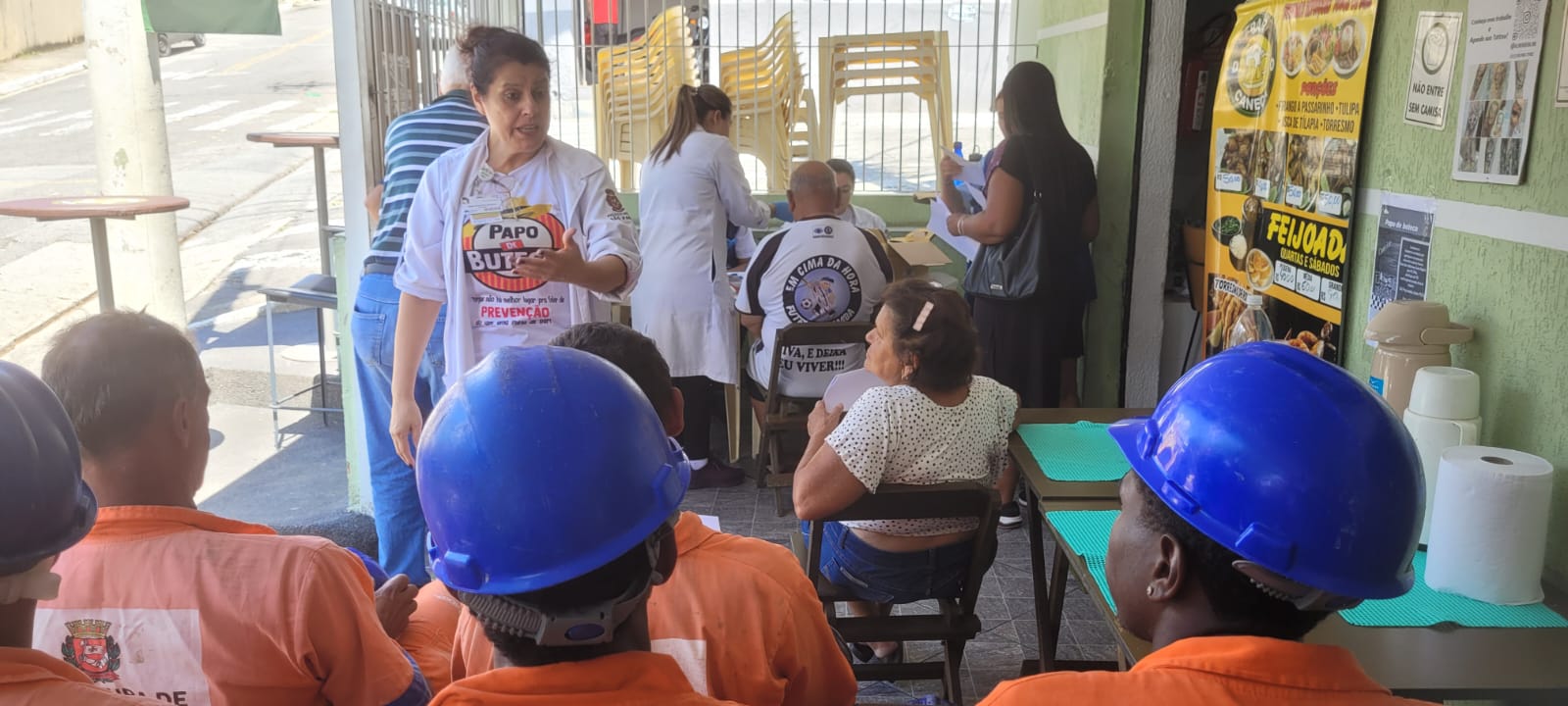 Na imagem, a enfermeira Mabel, uma mulher branca, de cabelo castanho e preso, usa jaleco branco e uma camiseta escrito 'Papo de Buteco'. Ela está palestrando para um grupo de homens que usam uniforme laranja e capacete azul. Todos estão de costas para a foto. No ambiente há uma idosa e equipe profissional da UBS